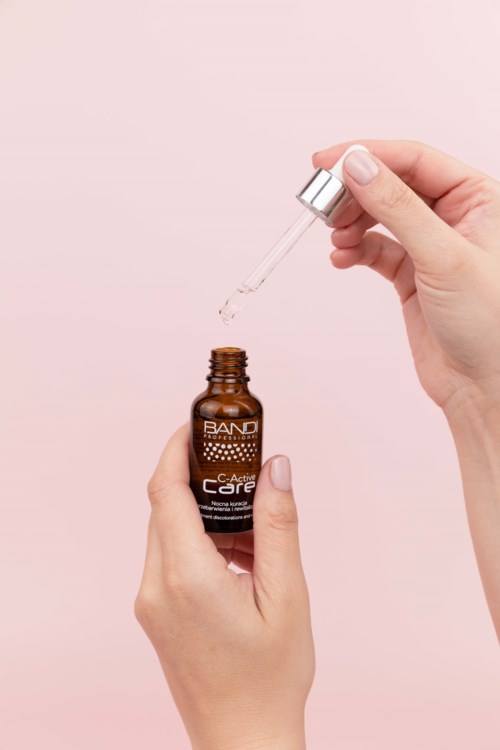 Eine Person hält eine braune Flasche mit Hautpflege-Serum, mit einem Tropfer, um Flüssigkeit zu verabreichen. Der Hintergrund ist hellrosa.