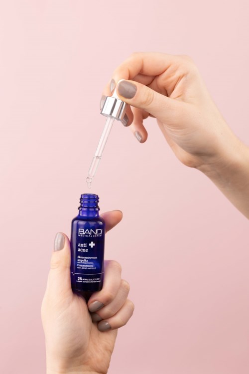 Eine Person mit schön lackierten Nägeln hält eine blaue Flasche mit Hautpflege-Serum und benutzt eine Pipette vor einem pinken Hintergrund.