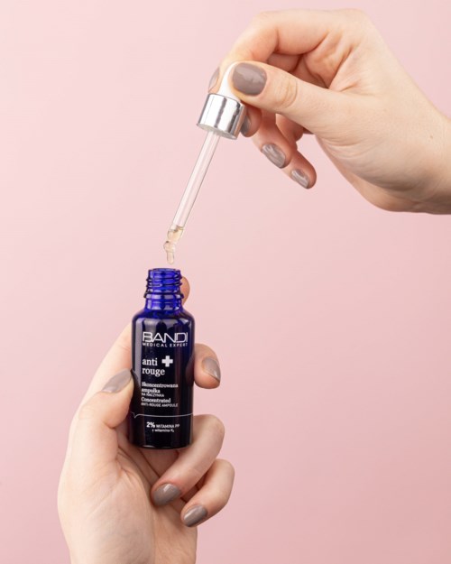En person med målade naglar använder en dropper för att applicera serum från en blå flaska mot en rosa bakgrund.