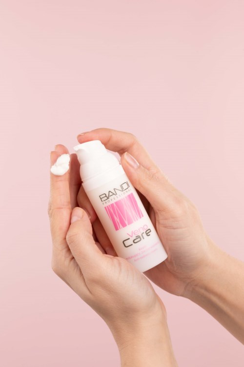 En person holder en hvit flaske med rosa og svart tekst, og doserer krem på fingeren, mot en rosa bakgrunn.