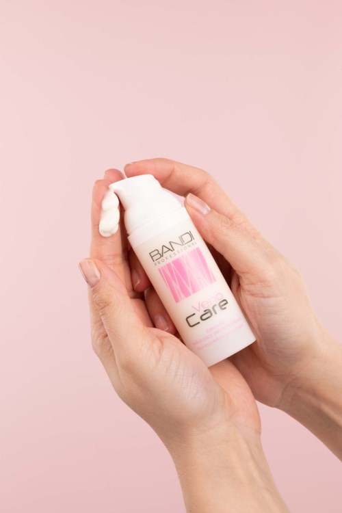 En hand som pressar lotion från en vit flaska med rosa etikett mot en rosa bakgrund.