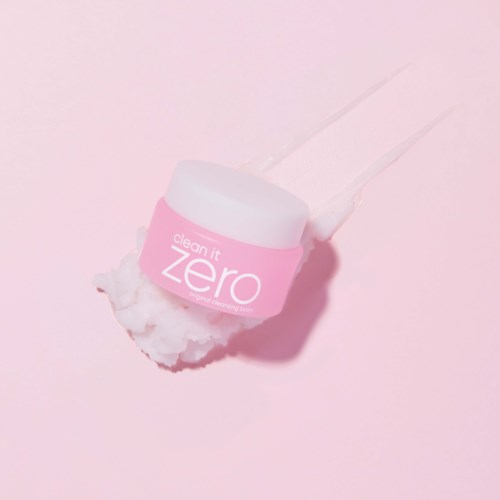 En lyserød beholder mærket 'clean it zero' med en vatpind og creme omkring den på en lyserød baggrund.