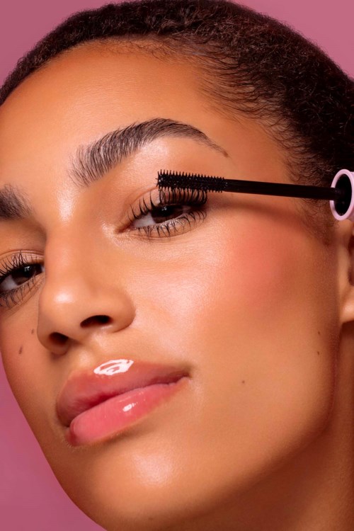A woman applying mascara on her eyelashes with a pink background.