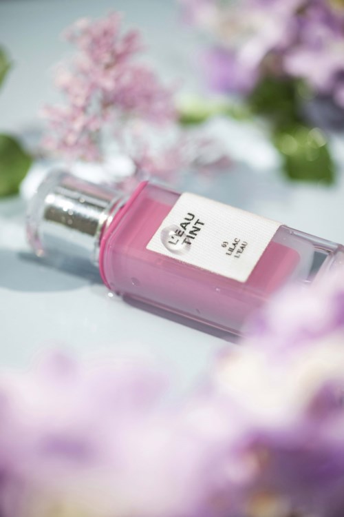 En rosa parfymeflaske merket "Eau Tain" ligger blant rosa og lilla blomster.