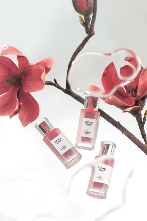 Three bottles of pink perfume hang from a branch with pink flowers, reflected on a glossy surface.