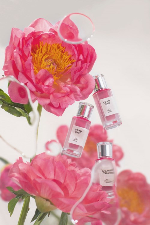 Three small pink perfume bottles hang among pink peonies with water drops, creating a soft, floral scene.