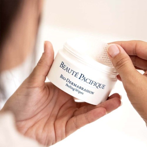 A person holding a white jar labeled 'BEAUTE PACIFIQUE' with peeling wipes inside.