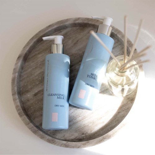 Twee lichtblauwe flessen huidverzorgingsproducten op een ronde houten schaal, één met pomp. Een reed diffuser met stokjes ligt ernaast.