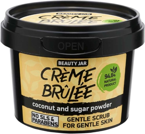 En svart burk med etiketten 'CRÈME BRÛLÉE' med kokos- och sockerpulver, mild skrubb och naturliga ingredienser vid 94,6%.
