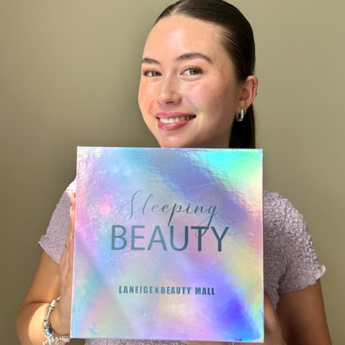 En kvinna ler och håller en holografisk box med texten 'Sleeping Beauty' från LaneigexBeauty Mall.