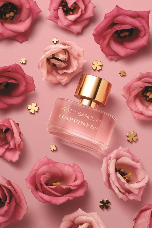 En parfume flaske kaldet 'Happiness' omgivet af pink blomster og små guld stjerner på en lyserød baggrund.