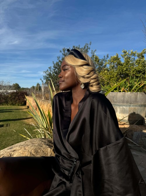 En kvinna med blond hår bär en svart rock, sitter utomhus med gröna växter och blå himmel bakom henne.