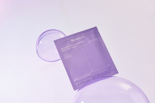 Violetti ihonhoitotuote pussi ja läpinäkyvä kasvomaski, ympärillä kirkkaat lasipallot vaalealla taustalla.