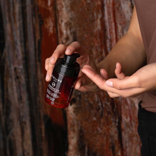 A person is holding a red bottle with a black lid, applying a product to their hand against a wooden background.