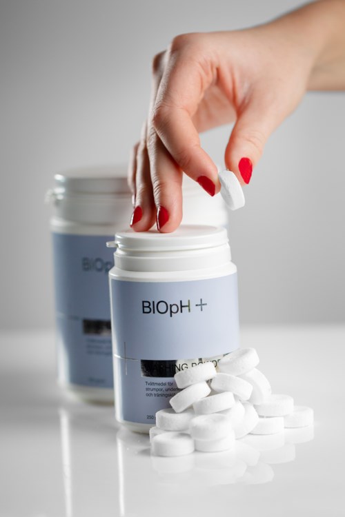 En hand tar en tablett från en behållare märkt "BIOpH+" med fler tabletter i närheten.