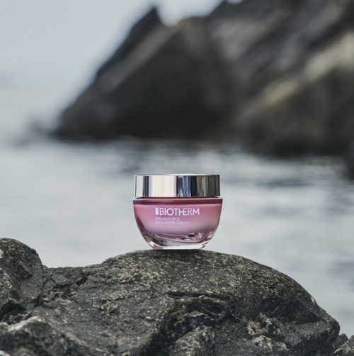 A pink jar of Biotherm skincare is placed on a rock near water, with rocks and water in the background.