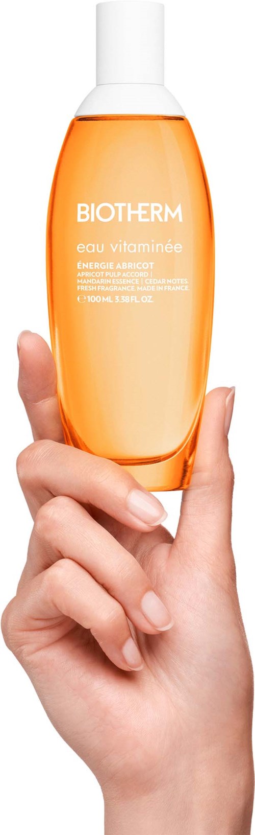 En hånd holder en oransje flaske med hvit lokk fra Biotherm, med tydelig tekst på etiketten, mot hvit bakgrunn.