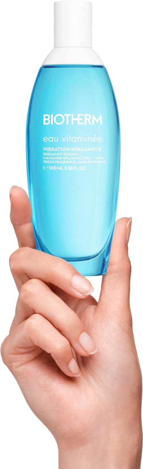 En hånd holder en blå flaske med Biotherm Eau Vitaminée, et fuktighetsgivende hudprodukt, mot en hvit bakgrunn.