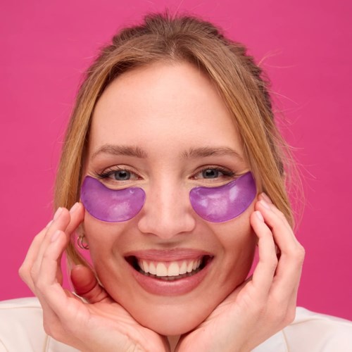 En kvinna ler och bär lila underögonmasker mot en rosa bakgrund.