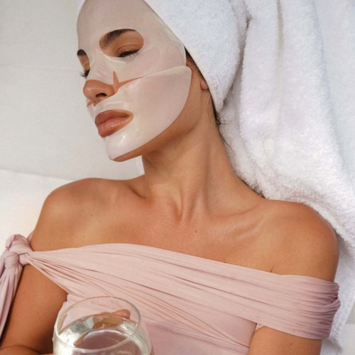 Frau mit Gesichtsmaske, Handtuch um den Kopf, trägt eine rosa Top, entspannt im Spa oder Salon.