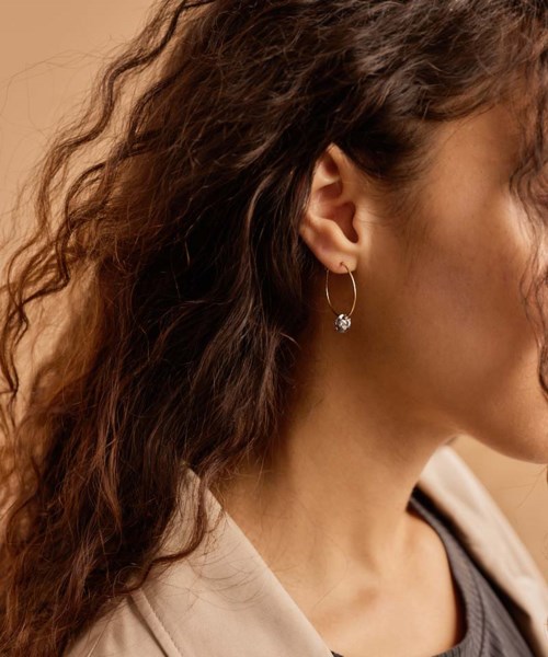 A woman wears hoop earrings with a dangling gemstone, with curly hair and a beige jacket.