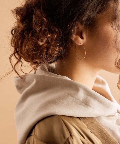 Close-up van een vrouw met krullend haar, grote oorringen en hoodie, zij aanzicht, neutrale achtergrond.