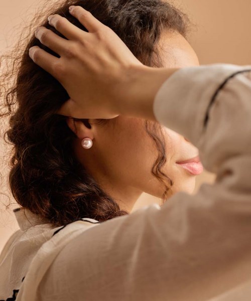 Een vrouw met krullend haar draagt parels, raakt haar hoofd zachtjes aan, met een neutrale achtergrond.