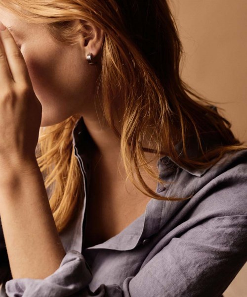 A woman with red hair partially covers her face with her hand, wearing a gray shirt and hoop earrings.