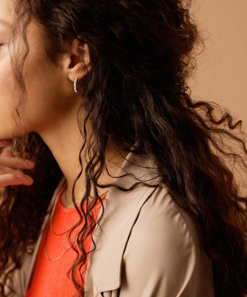 A woman with dark, curly hair wearing earrings and a beige jacket over an orange top.