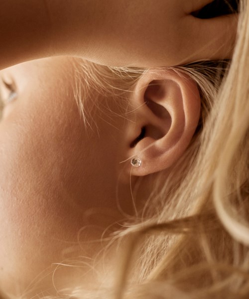 Close-up of a person's ear with a small earring, and blonde hair flowing nearby, showing soft skin and delicate features.