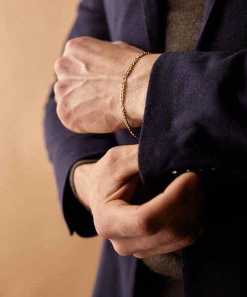A person wears a dark blazer with a gold bracelet, holding their arm against their chest. The background is plain.