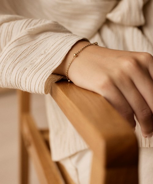 Eine Person trägt ein goldenes Armband und hält die Hand auf eine Holzwand. Sie trägt ein cremefarbenes Langarmhemd.