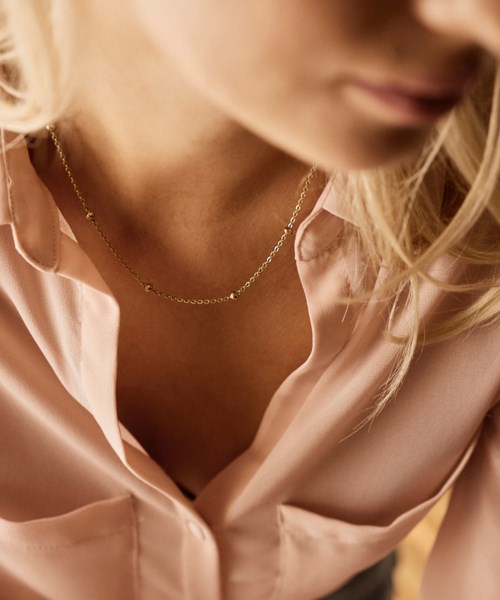 Eine Frau trägt ein hellrosa Hemd und eine kleine Goldkette, das Gesicht ist teilweise sichtbar und hat blondes Haar.