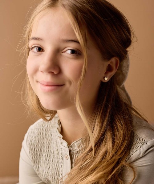 A young girl with long, wavy hair, smiling softly, wearing a light-colored blouse, and sitting against a beige background.