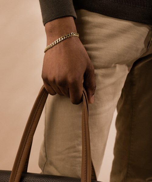 A person holds a brown bag, wearing a gold chain bracelet, beige pants, and a grey top.