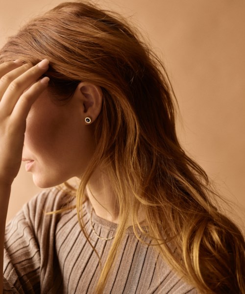 A woman with long, wavy red hair, wearing a striped beige and brown top, touches her hair with her hand.