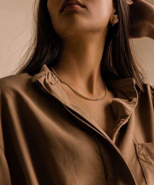 Eine Frau mit langen dunklen Haaren, trägt eine braune seidige Bluse und eine Goldkette, posiert elegant.