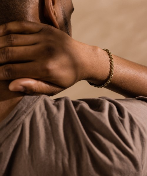 A person touches their neck with one hand, wears a bracelet, and a brown shirt. The background is plain.