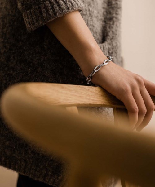 Die Hand einer Person mit einem Silberarmband ruht auf einer Holzfläche, getragen wird ein gemütlicher Pullover.