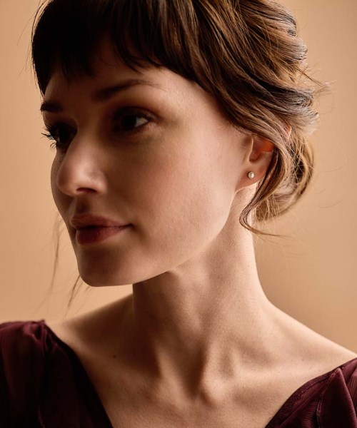 A woman with short brown hair and small earrings, looking to the side, in soft lighting.