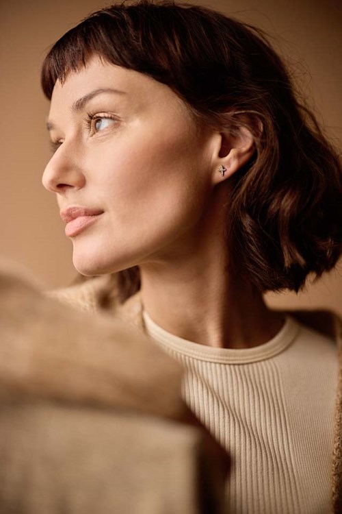 A woman with short, dark hair looks to the side, wearing a beige top and pearl earrings.