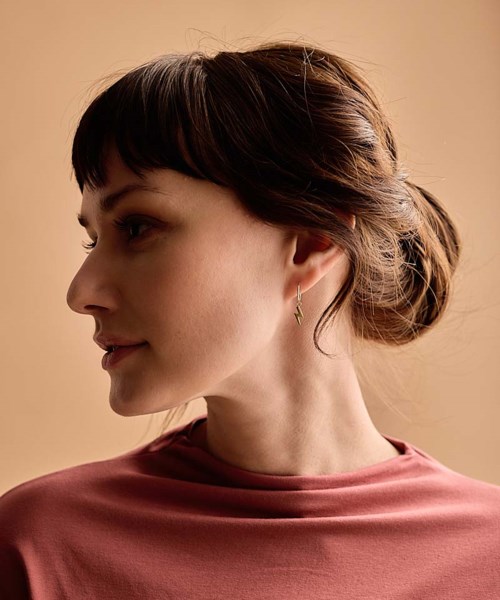 A woman with brown hair in a bun, wearing a rose-colored top and small earrings, looking to the left against a beige background.