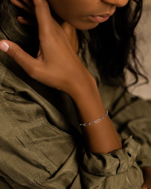 A person with dark curly hair is resting her hand on her shoulder, wearing a silver bracelet and a loose olive-green garment.