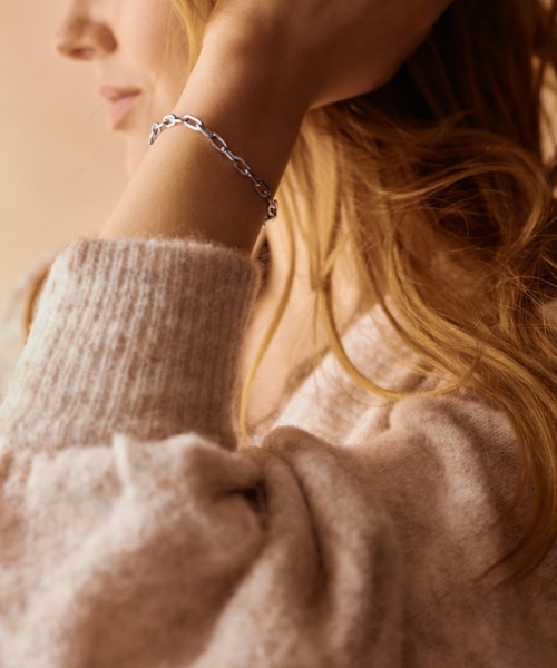 A woman with red hair wearing a silver bracelet and a beige sweater, with her hand near her face.