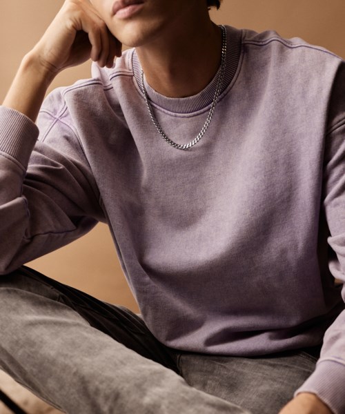 Ein junger Mann trägt ein lavendelblaues Sweatshirt, silberne Halskette und sitzt mit der Hand am Gesicht vor einem beigen Hintergrund.