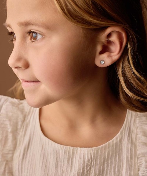 Close-up of a young girl with light skin and blonde hair, wearing small stud earrings and a cream-colored top.