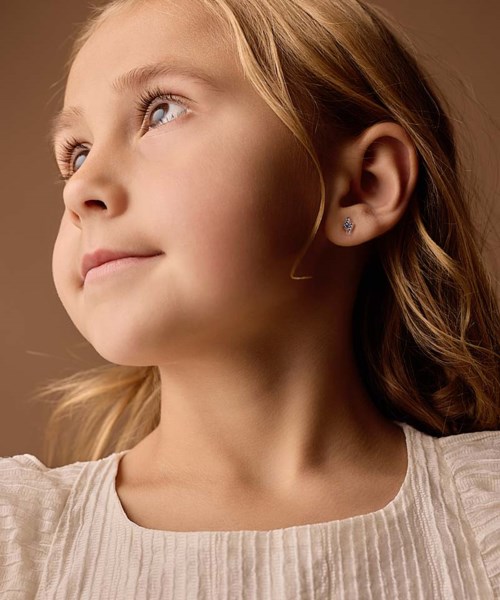 Young girl with light brown hair, looking upwards with a gentle smile, wearing a white top and small earrings.