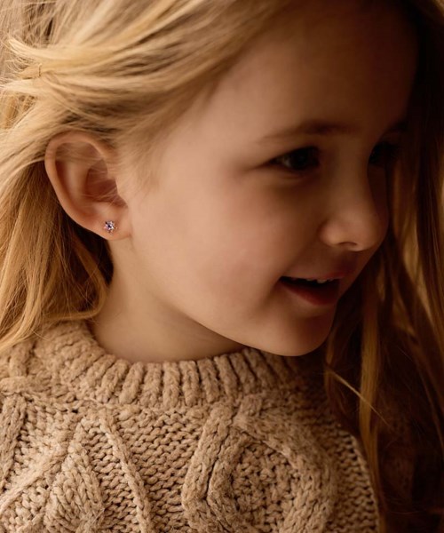 A young girl with blonde hair wearing small earrings and a beige knit sweater, smiling and looking to the side.