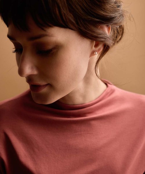 A woman with short brown hair and earrings wearing a pink top, looking down gently.