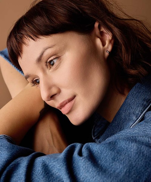 Close-up of a young woman with dark hair and a gentle smile resting her head on her arm, wearing a denim jacket.